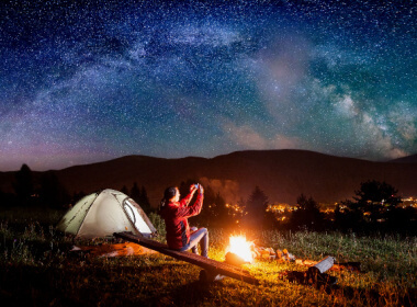 a woman camping out under a night sky filled with satrs. She has a camp fire going.